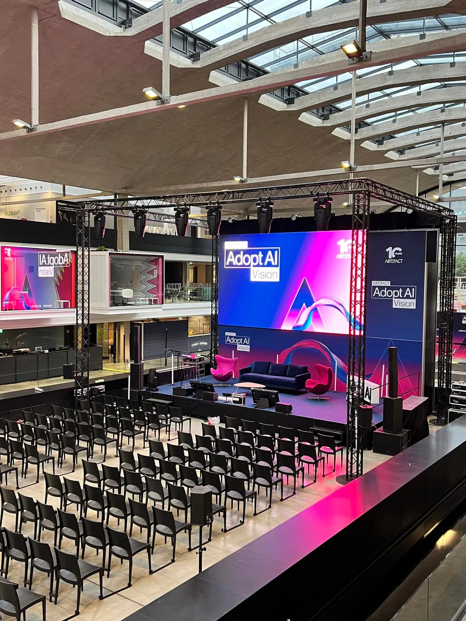 Plénière sous verrière avec scène sous structure lumière, grand écran LED, canapé et fauteuils d'intervenants et rangées de chaises noires