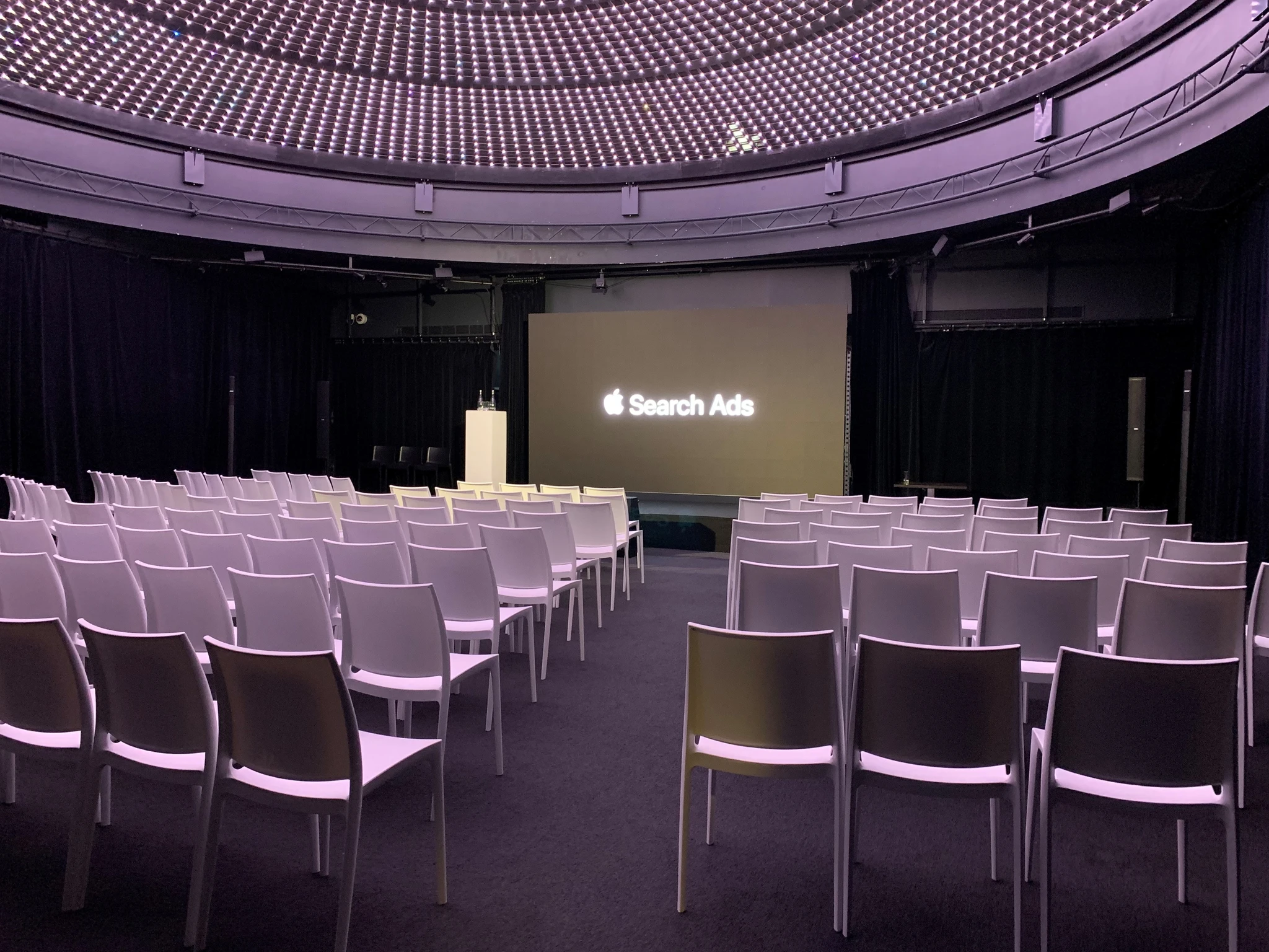 Salle de conférence circulaire sous coupole lumineuse, chaises coques blanches en théâtre avec allée centrale, pupitre blanc et écran de projection