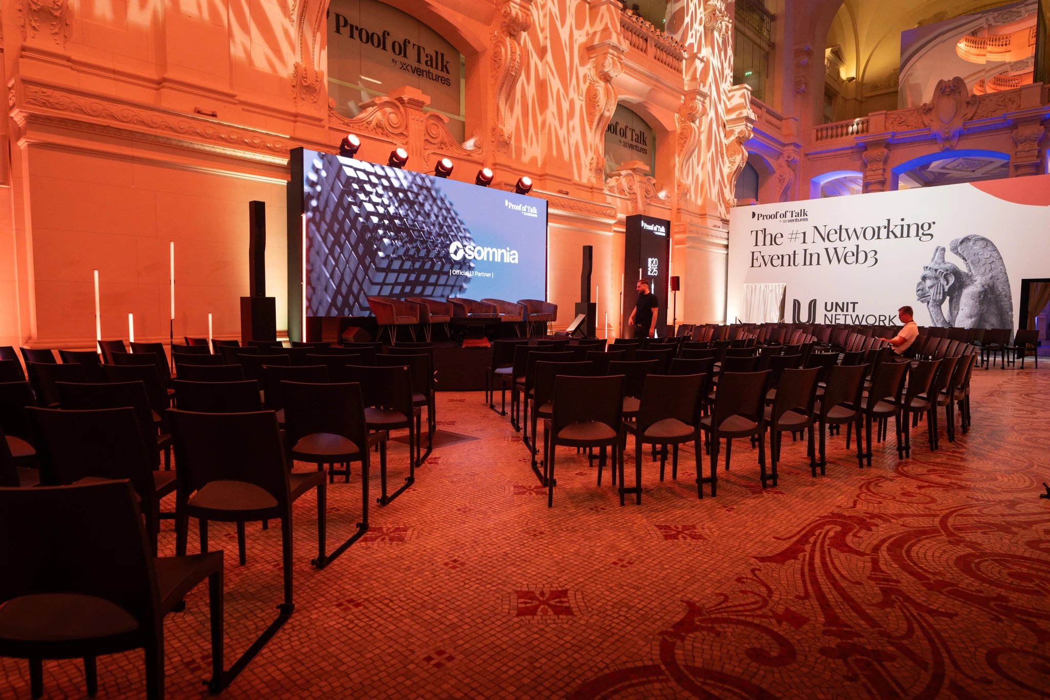 Conférence dans un hall historique éclairé en orange, avec écran LED sur scène, fauteuils d'intervenants et rangées de chaises noires