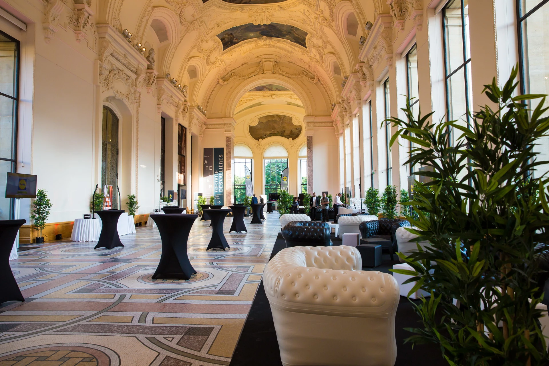 Galerie d'un palais parisien aménagée en lounge avec fauteuils Chesterfield blancs et noirs, mange-debout en housse noire et plantes vertes