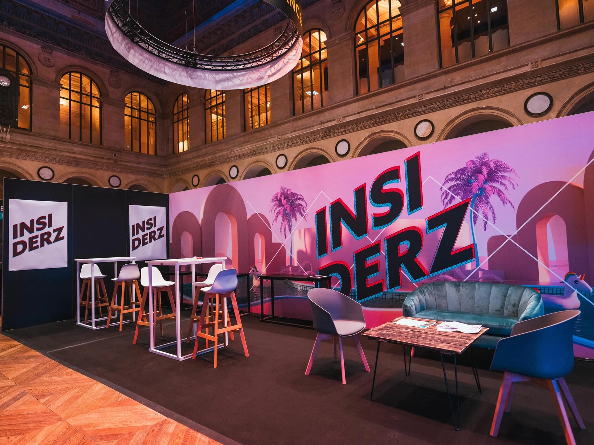 Espace networking dans un hall historique avec mange-debout blancs, tabourets hauts en bois, canapé en velours et grand fond imprimé rose