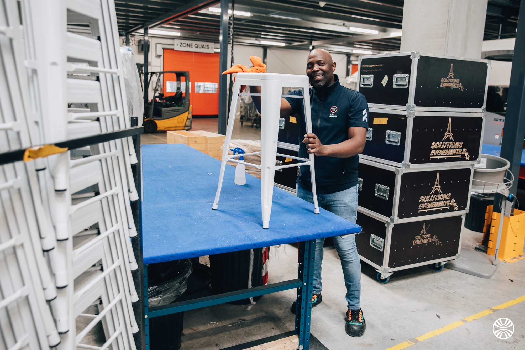 Préparateur nettoyant un tabouret en métal blanc dans l'entrepôt Solutions Événements, à côté de flight cases